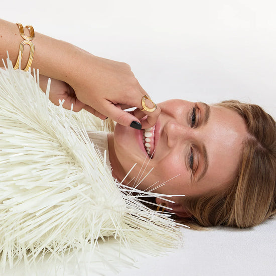 A close-up lifestyle shot of a smiling woman lying down, wearing gold rings and a gold C-shaped bracelet, with a fluffy white faux-fur collar around her neck against a clean white background.