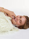 A close-up lifestyle shot of a smiling woman lying down, wearing gold rings and a gold C-shaped bracelet, with a fluffy white faux-fur collar around her neck against a clean white background.