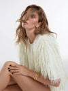 A woman wearing a cream-colored fringed top and gold jewelry, posing against a plain white studio background.