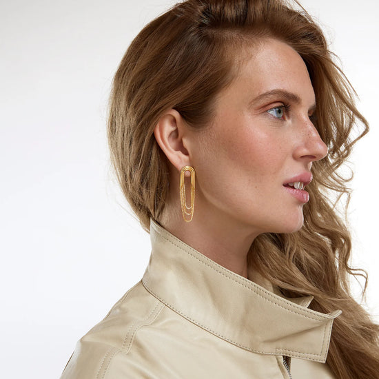 A woman in profile wearing a beige leather jacket and a gold-toned geometric hoop earring, photographed against a clean white studio background.