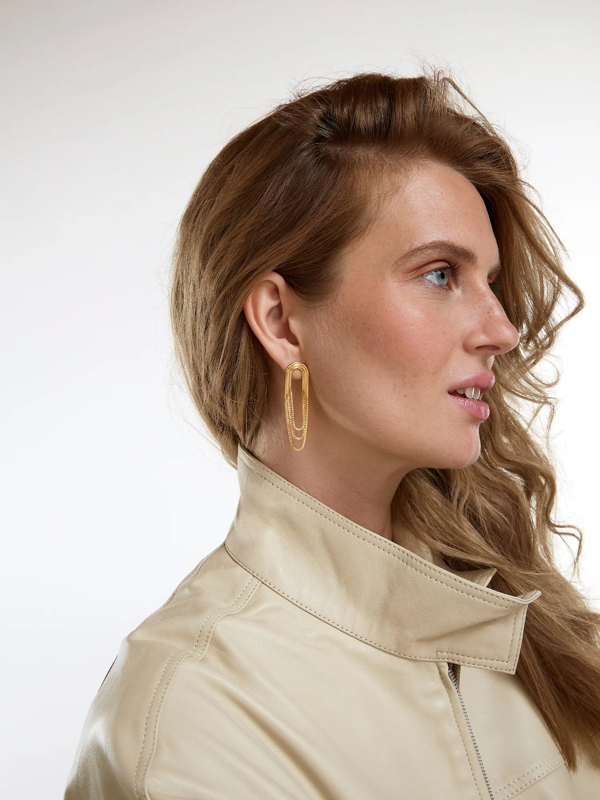 A woman in profile wearing a beige leather jacket and a gold-toned geometric hoop earring, photographed against a clean white studio background.