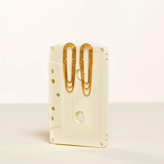 A cream-colored cassette tape-shaped desk organizer holding two gold paperclips, set against a clean off-white background.