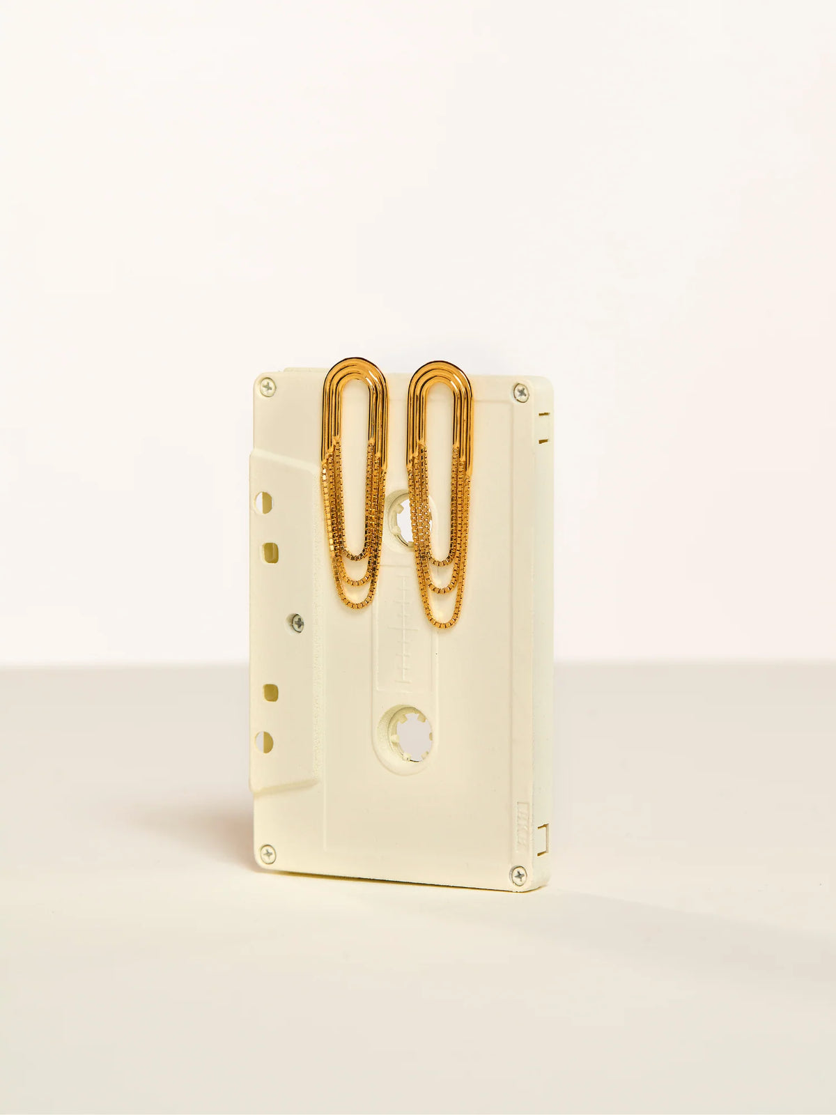 A cream-colored cassette tape-shaped desk organizer holding two gold paperclips, set against a clean off-white background.