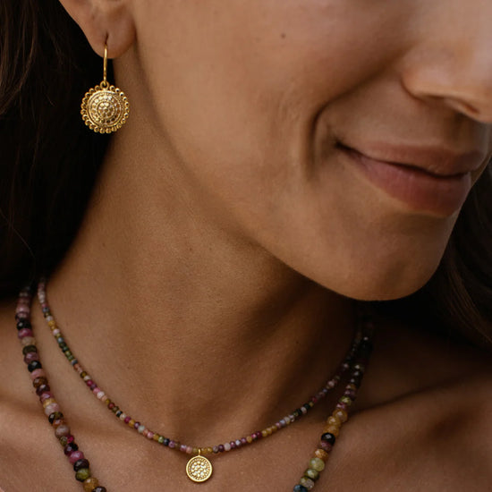 Close-up of a person wearing gold earrings and a multicoloured beaded necklace from Anna Beck