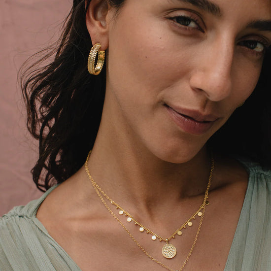 Woman wearing gold hoop earrings and necklaces against a neutral background from Anna Beck