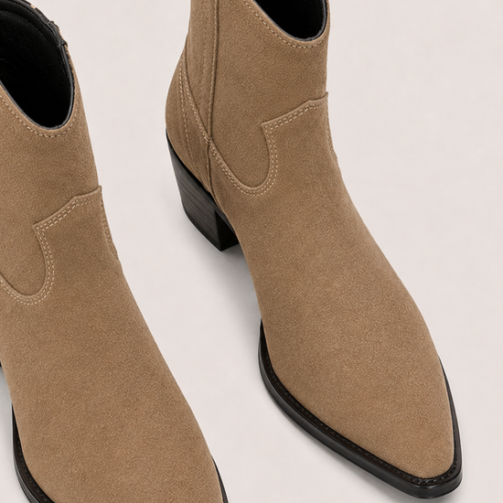 A pair of pointed-toe ankle boots in tan suede with black soles and low block heels, shown on a light beige background.