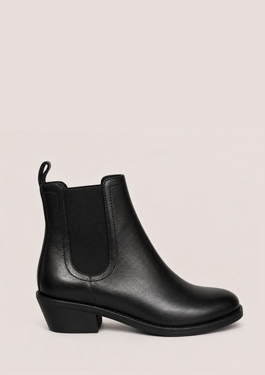 A single black leather Chelsea-style ankle boot with a low block heel and elastic side gore, photographed on a plain light warm-gray background.