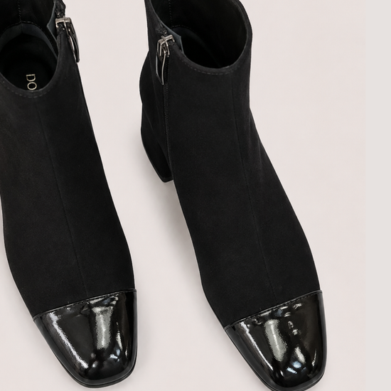 A pair of black ankle boots with a glossy patent leather cap toe, shown from a top-down angle on a light beige background.