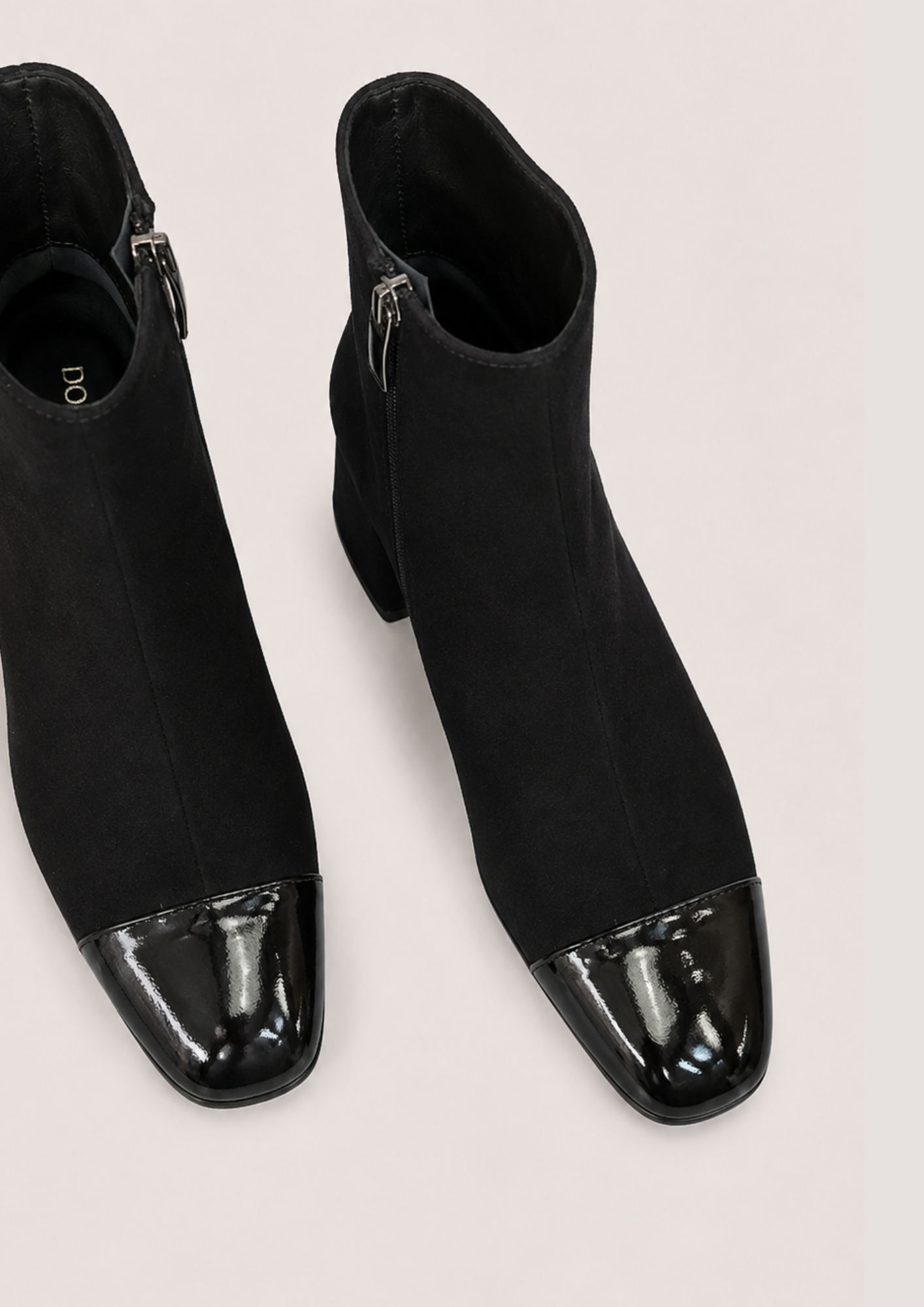 A pair of black ankle boots with a glossy patent leather cap toe, shown from a top-down angle on a light beige background.