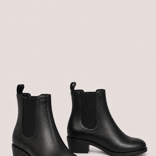 A pair of black leather Chelsea ankle boots with elastic side panels and low block heels, displayed against a solid pale pink background.