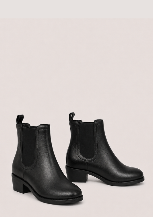A pair of black leather Chelsea ankle boots with elastic side panels and low block heels, displayed against a solid pale pink background.