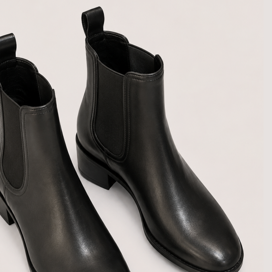 A pair of black leather Chelsea-style ankle boots with elastic side panels and low block heels, photographed on a plain light beige studio background.