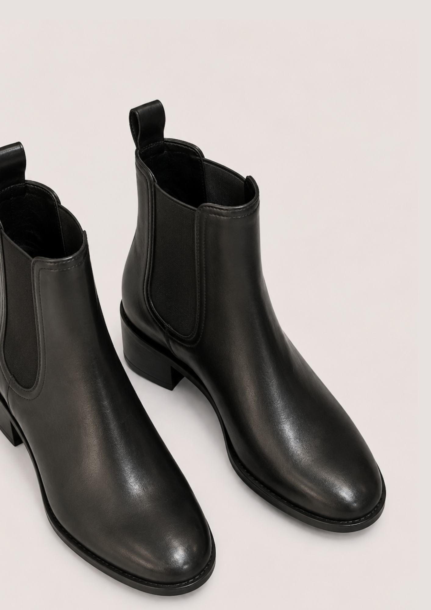 A pair of black leather Chelsea-style ankle boots with elastic side panels and low block heels, photographed on a plain light beige studio background.