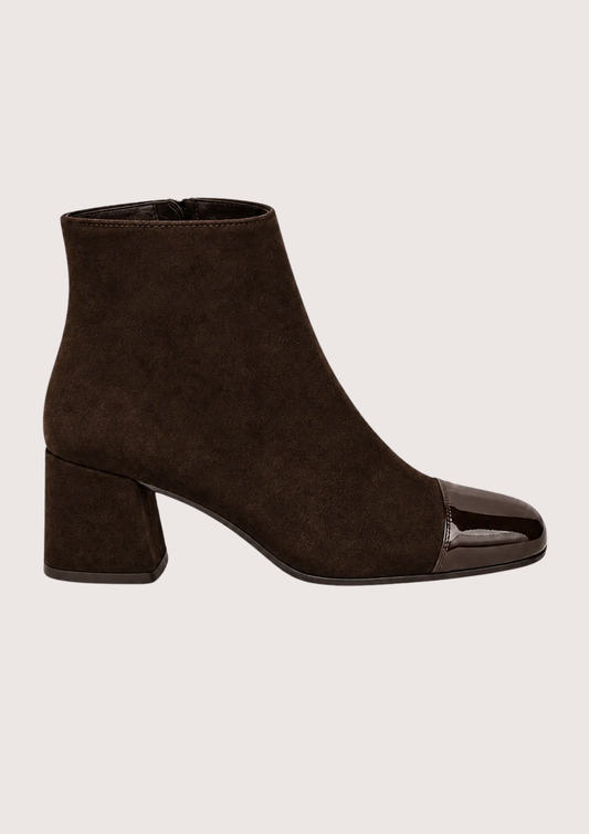 A dark brown suede ankle boot with a glossy black patent leather toe cap and a low block heel, shown in profile against a light beige background.