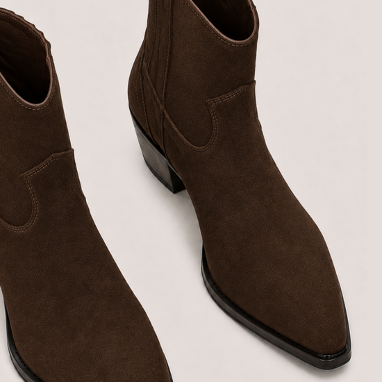 A pair of dark brown suede ankle boots with pointed toes and low block heels, shown from a high-angle perspective on a light gray background.