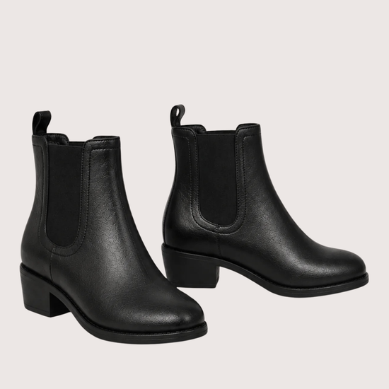 A pair of black leather Chelsea ankle boots with elastic side panels and low block heels, displayed against a solid pale pink background.