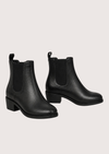 A pair of black leather Chelsea ankle boots with elastic side panels and low block heels, displayed against a solid pale pink background.