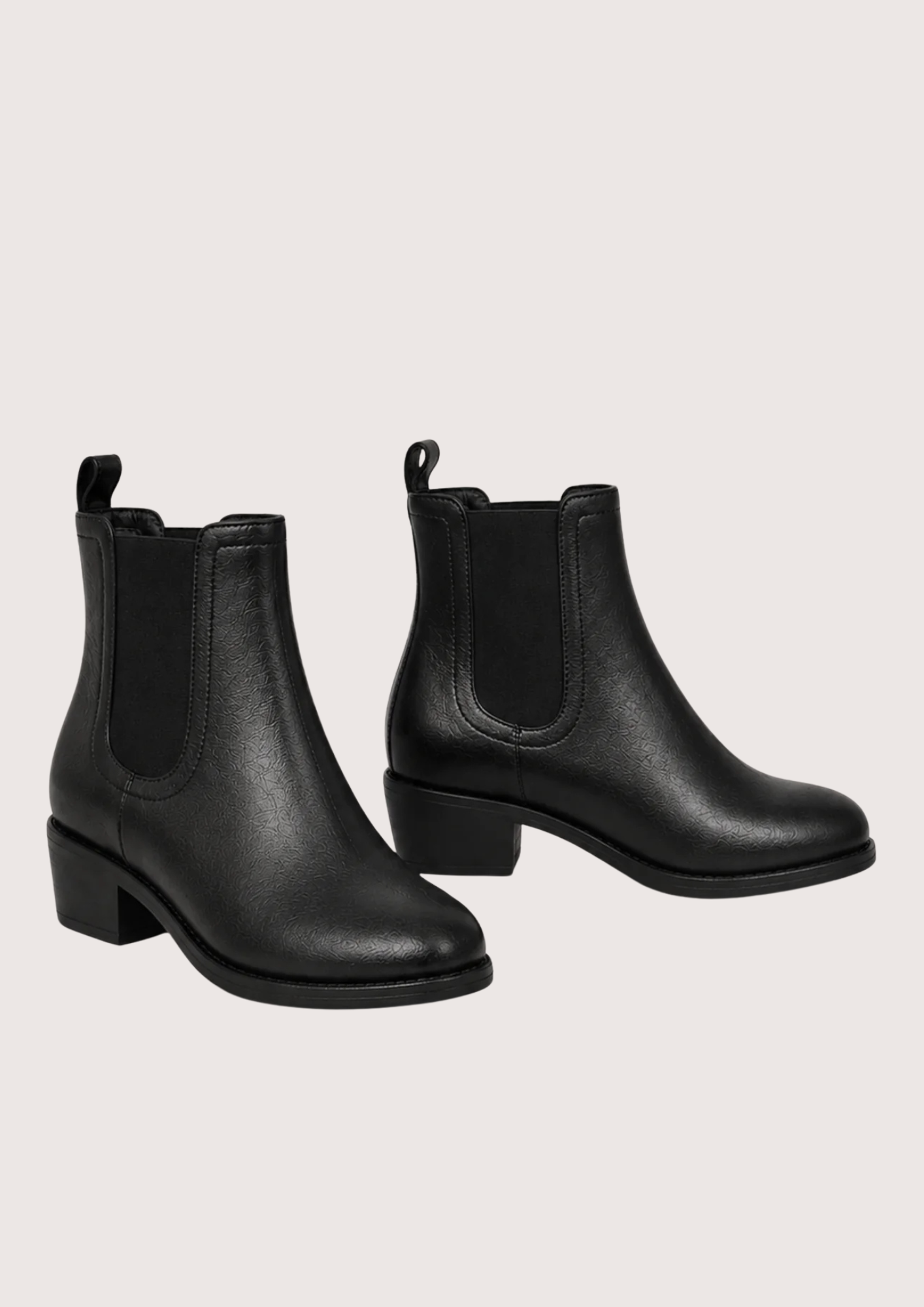 A pair of black leather Chelsea ankle boots with elastic side panels and low block heels, displayed against a solid pale pink background.