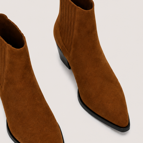 A pair of pointed-toe ankle boots in brown suede with black low heels, shown from an elevated angle on a light beige background.