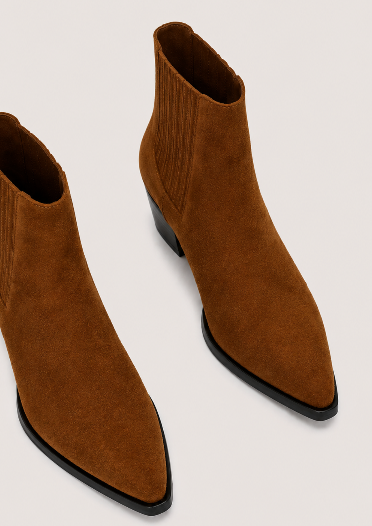 A pair of pointed-toe ankle boots in brown suede with black low heels, shown from an elevated angle on a light beige background.