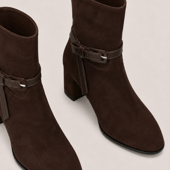 A pair of dark brown suede ankle boots with a block heel and a matching faux-leather strap detail on the side, displayed on a light beige background.
