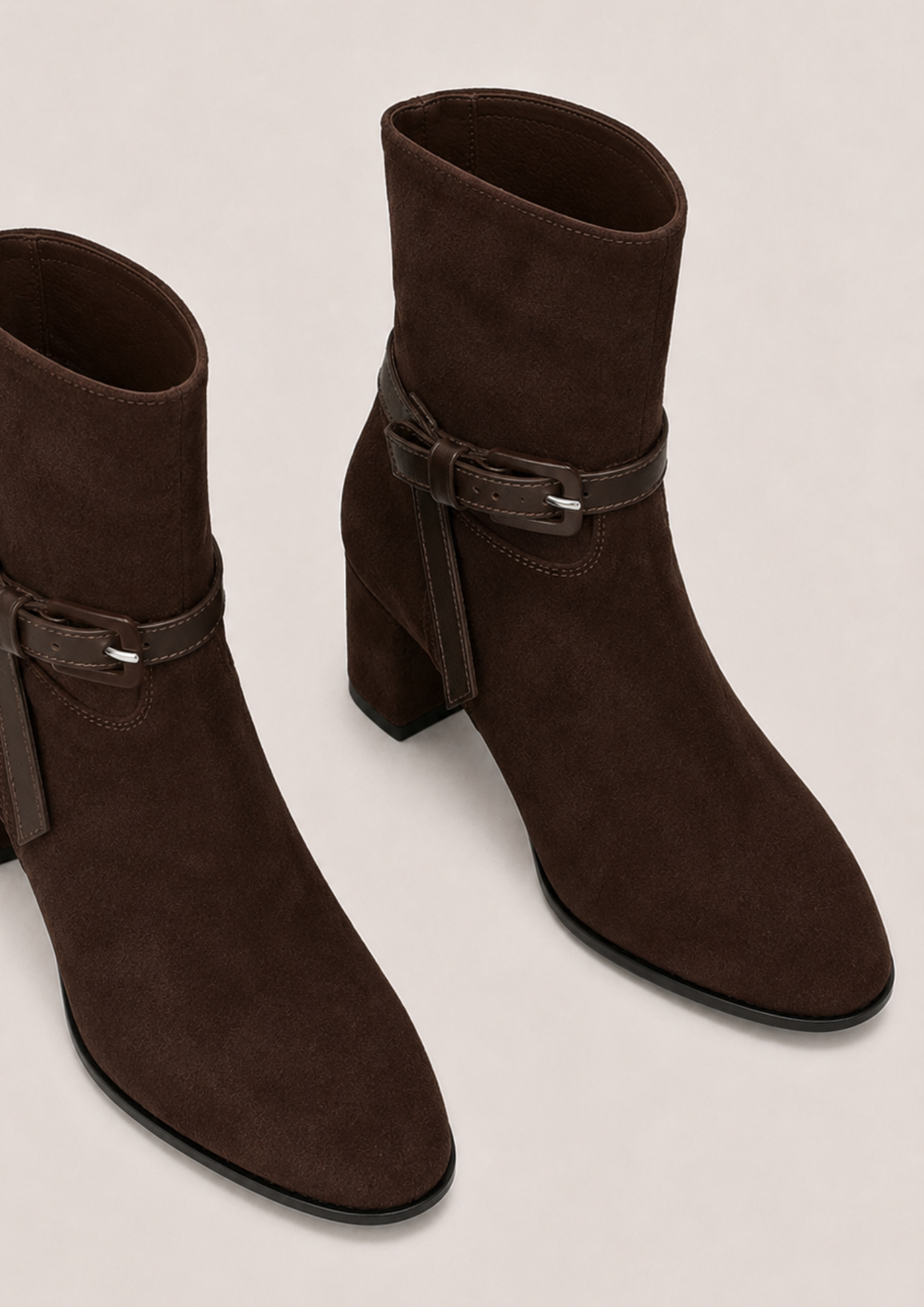A pair of dark brown suede ankle boots with a block heel and a matching faux-leather strap detail on the side, displayed on a light beige background.