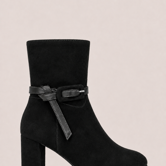 A black suede ankle boot with a block heel and a decorative black strap with a silver buckle detail around the ankle, shown against a solid light beige background.