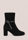 A black suede ankle boot with a block heel and a decorative black strap with a silver buckle detail around the ankle, shown against a solid light beige background.