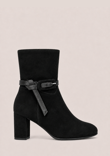 A black suede ankle boot with a block heel and a decorative black strap with a silver buckle detail around the ankle, shown against a solid light beige background.