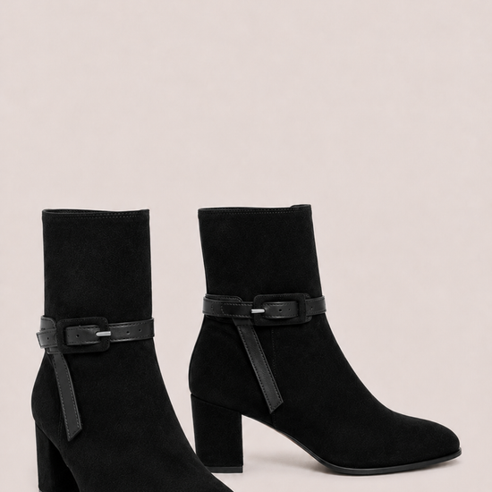 A pair of black suede ankle boots with block heels and a matching black faux-leather strap detail across the instep, displayed against a solid light beige background.