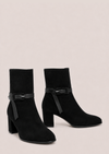 A pair of black suede ankle boots with block heels and a matching black faux-leather strap detail across the instep, displayed against a solid light beige background.