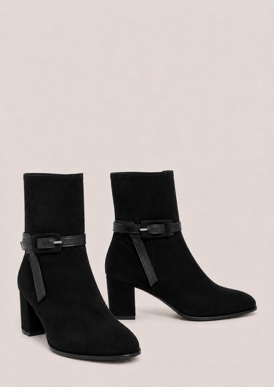 A pair of black suede ankle boots with block heels and a matching black faux-leather strap detail across the instep, displayed against a solid light beige background.