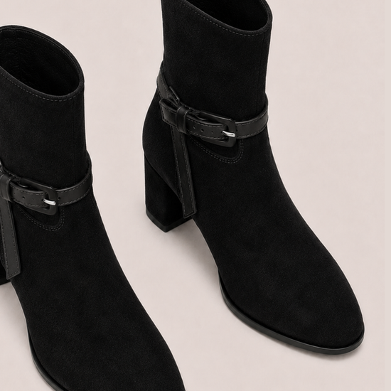 A pair of black suede ankle boots with a block heel and a decorative strap detail with a buckle on the outer side, shown against a light beige background.