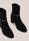 A pair of black suede ankle boots with a block heel and a decorative strap detail with a buckle on the outer side, shown against a light beige background.