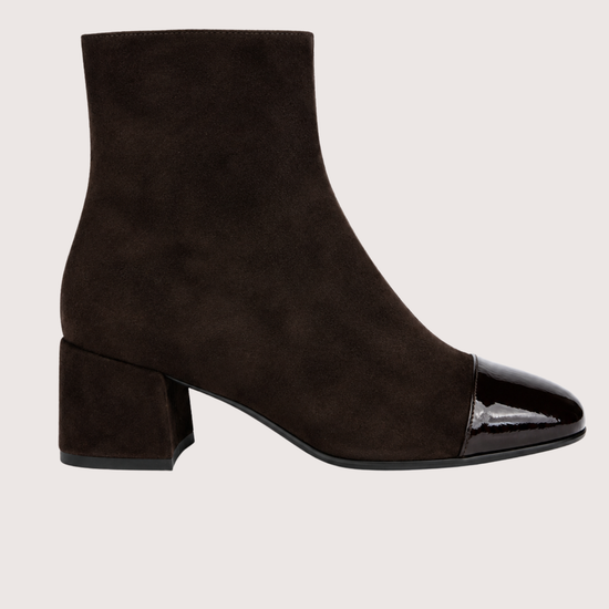 A dark brown suede ankle boot with a glossy black patent leather toe cap and a low block heel, shown in profile against a light beige background.
