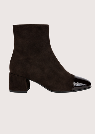 A dark brown suede ankle boot with a glossy black patent leather toe cap and a low block heel, shown in profile against a light beige background.