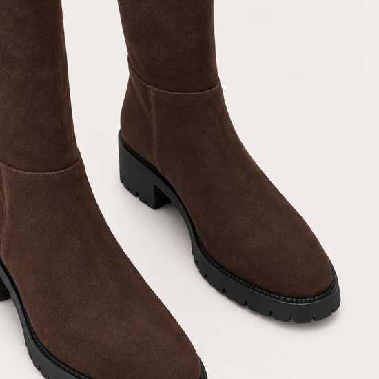 A pair of dark brown suede over-the-knee boots with chunky black block heels and lug soles, shown from a close-up angle against a light off-white background.
