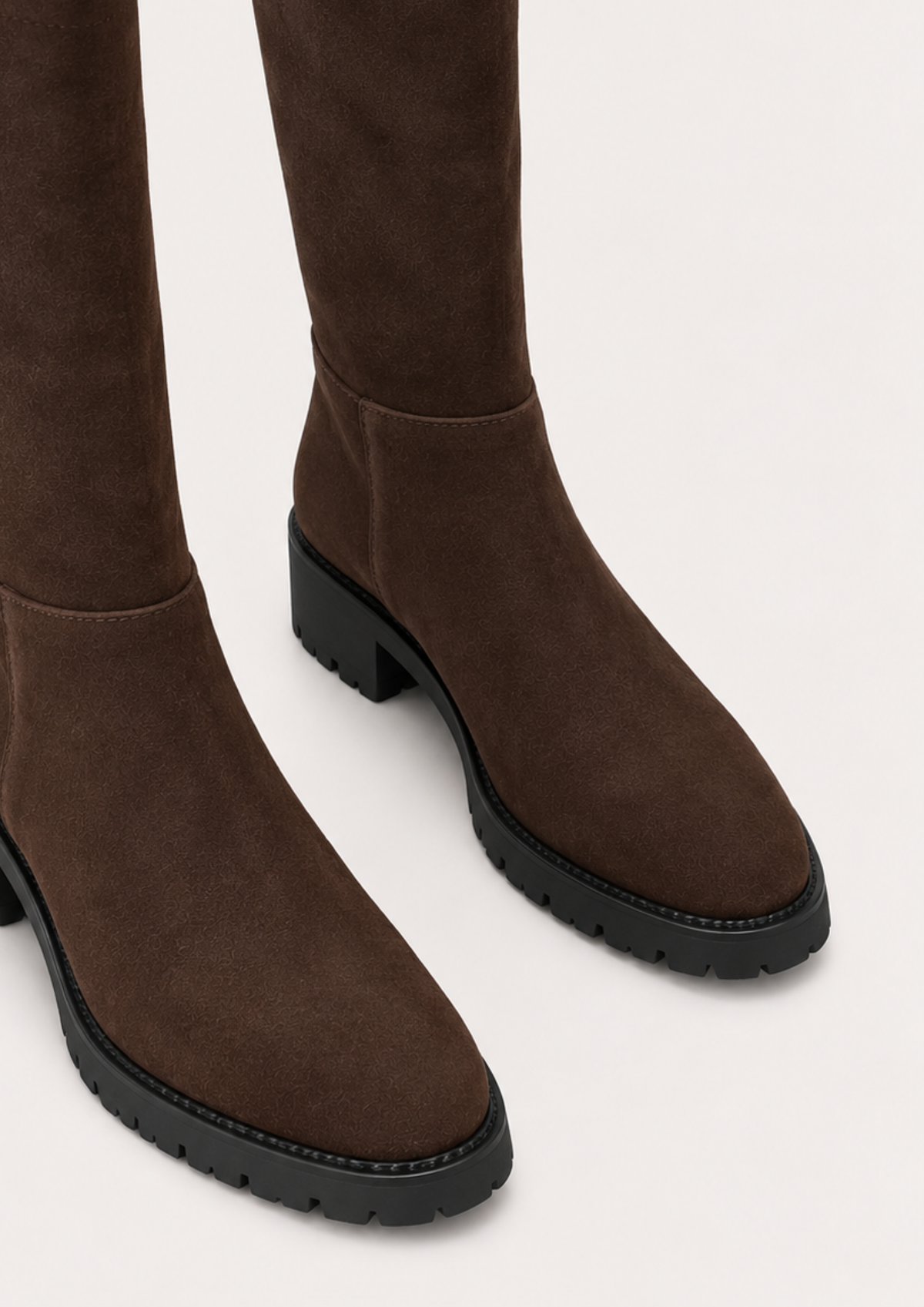 A pair of dark brown suede over-the-knee boots with chunky black block heels and lug soles, shown from a close-up angle against a light off-white background.