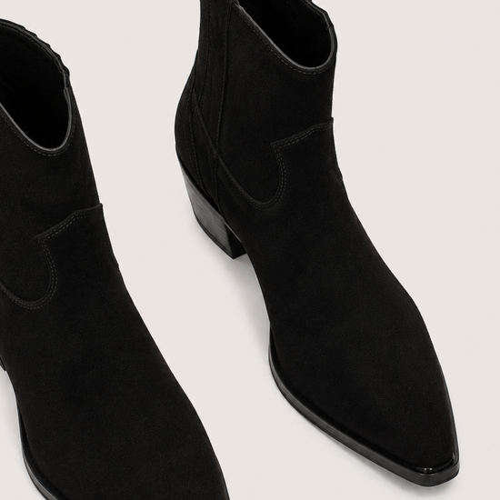 A pair of black suede ankle boots with pointed toes and low block heels, shown from a high-angle perspective on a light gray background.