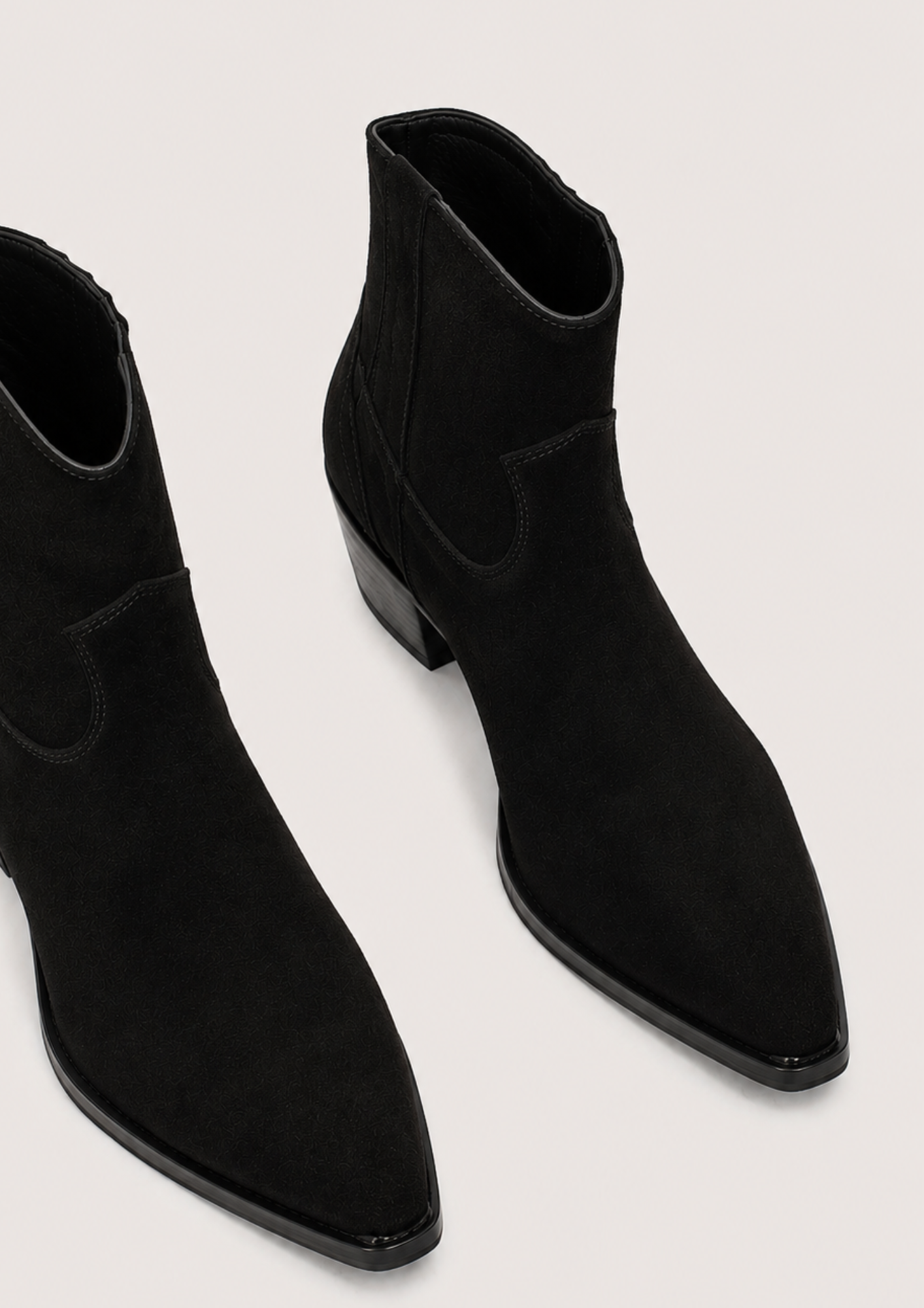 A pair of black suede ankle boots with pointed toes and low block heels, shown from a high-angle perspective on a light gray background.
