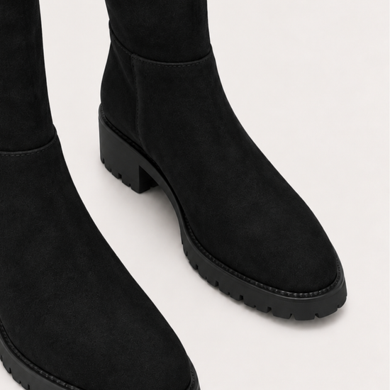 Black suede over-the-knee boots with chunky lug soles, shown from a close-up angle on a light gray background.