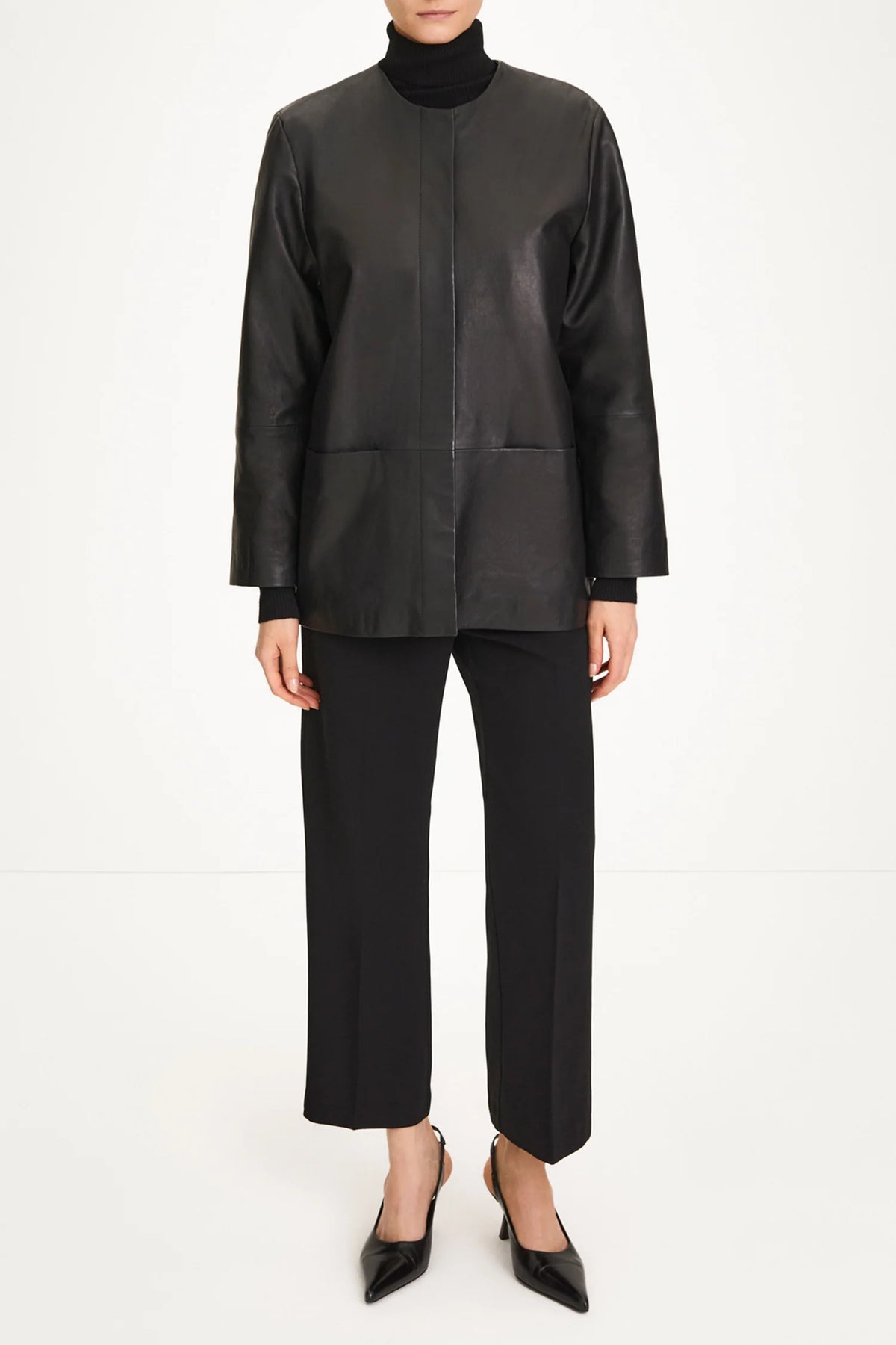 A model wearing a black leather jacket with a mock neck, black wide-leg cropped trousers, and black pointed-toe heels, photographed against a seamless white studio background.