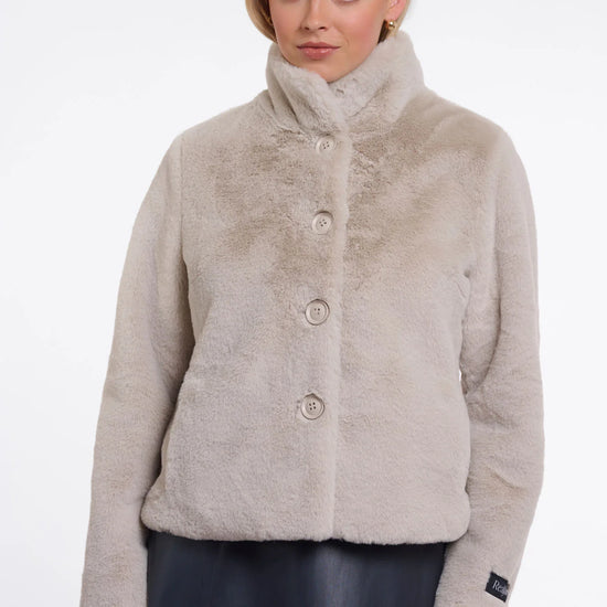 A woman wearing a light beige faux fur jacket with a high stand-up collar and three front buttons, paired with a dark skirt, against a plain white studio background.