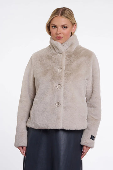A woman wearing a light beige faux fur jacket with a high stand-up collar and three front buttons, paired with a dark skirt, against a plain white studio background.