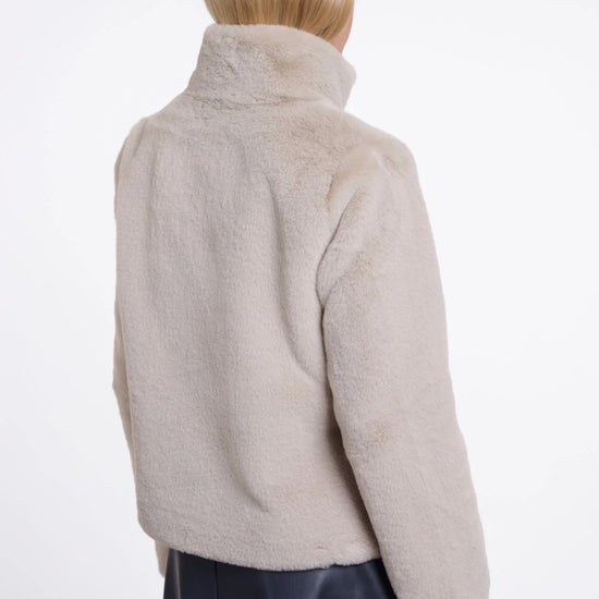 A model shown from the back wearing a light beige fuzzy cropped jacket with a high collar and a dark navy skirt, against a plain white studio background.