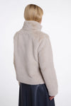 A model shown from the back wearing a light beige fuzzy cropped jacket with a high collar and a dark navy skirt, against a plain white studio background.
