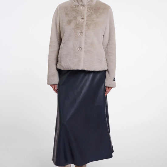 A model stands against a light gray studio background wearing a light beige cropped faux-fur jacket with a high collar and three front buttons, paired with a dark navy midi skirt and beige ankle boots.