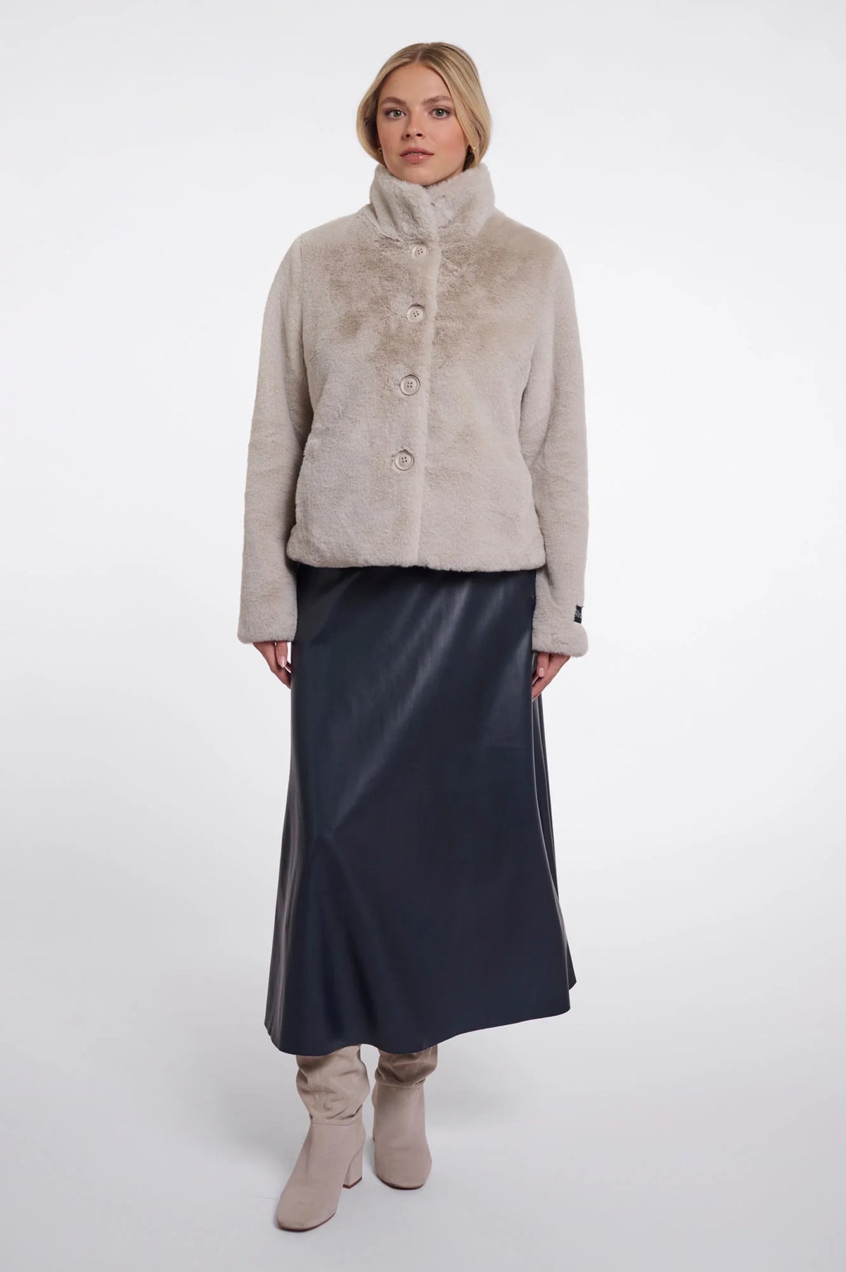 A model stands against a light gray studio background wearing a light beige cropped faux-fur jacket with a high collar and three front buttons, paired with a dark navy midi skirt and beige ankle boots.