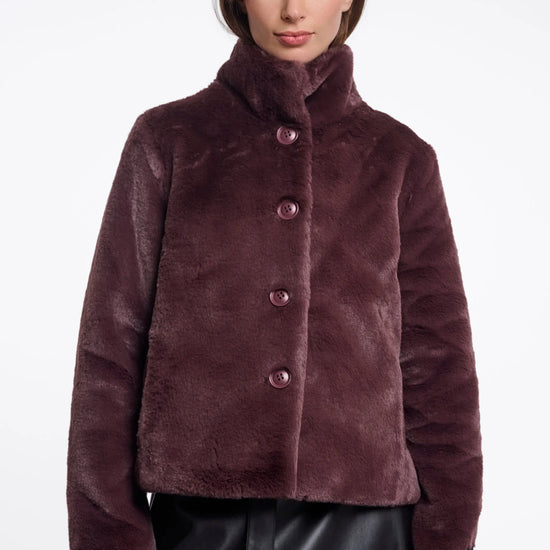 A woman wearing a plush, cropped faux fur jacket in a deep plum-brown color with matching buttons, paired with a black skirt, against a plain white studio background.