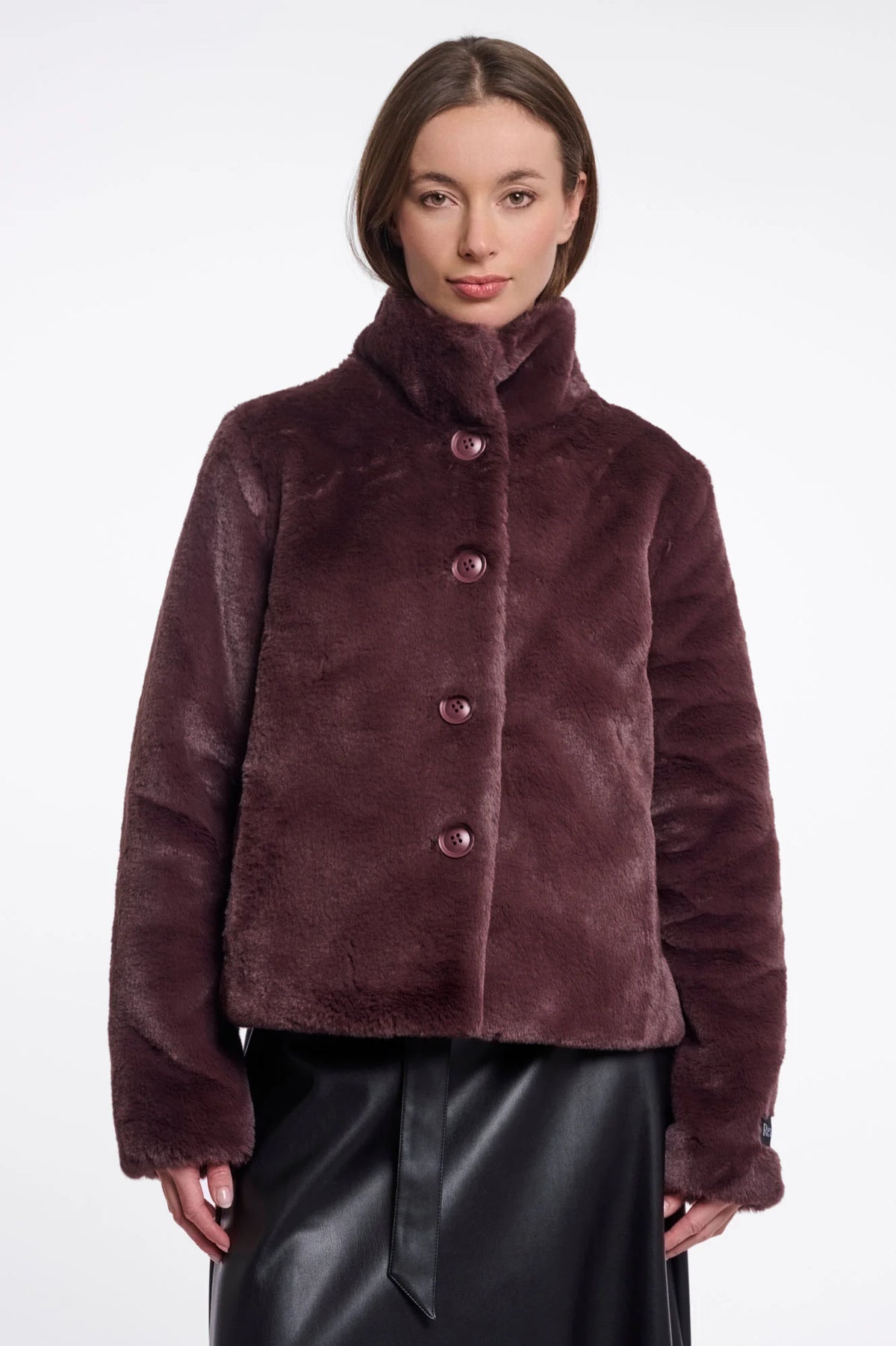 A woman wearing a plush, cropped faux fur jacket in a deep plum-brown color with matching buttons, paired with a black skirt, against a plain white studio background.